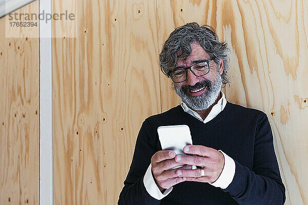 Smiling senior man text messaging through smart phone in front of wall