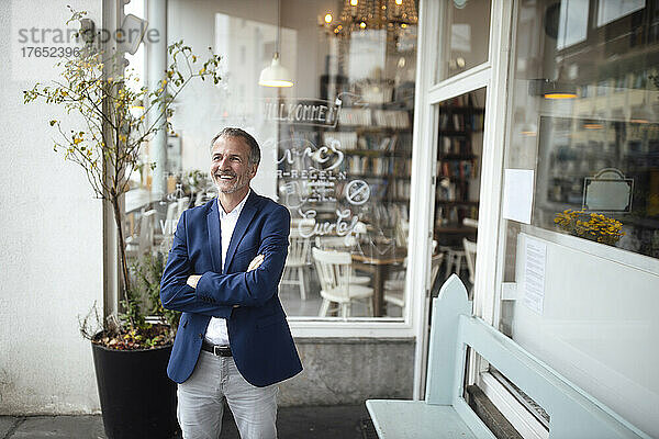 Happy businessman with arms crossed standing in front of glass window
