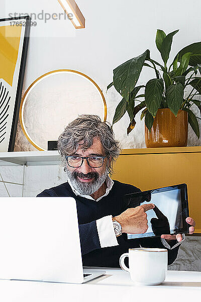 Lächelnder Freiberufler erklärt Tablet-PC per Videoanruf auf Laptop zu Hause