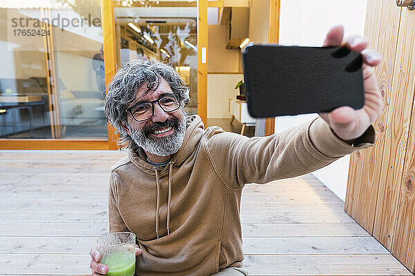 Lächelnder Mann mit einem Glas Smoothie  der ein Selfie mit dem Handy macht