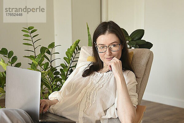 Lächelnde Frau mit Laptop sitzt zu Hause auf einem Stuhl