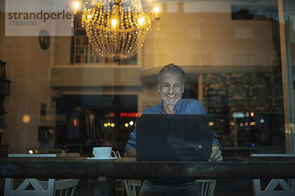 Lächelnder Freiberufler  der nachts im beleuchteten Café am Laptop arbeitet