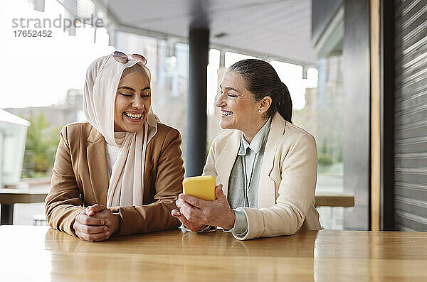 Glückliche Geschäftsfrau  die am Tisch im Café sitzt und ihr Smartphone teilt