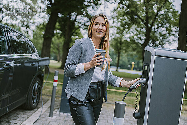 Frau mit Einwegbecher steht an der Ladestation für Elektrofahrzeuge