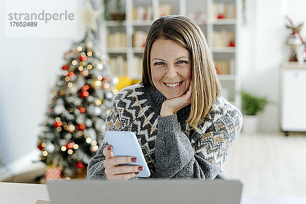 Glückliche Frau mit der Hand am Kinn und dem Mobiltelefon  die zu Hause im Wohnzimmer sitzt