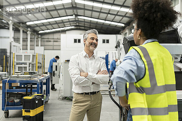 Lächelnder Geschäftsmann steht mit verschränkten Armen und diskutiert mit Ingenieur in der Fabrik