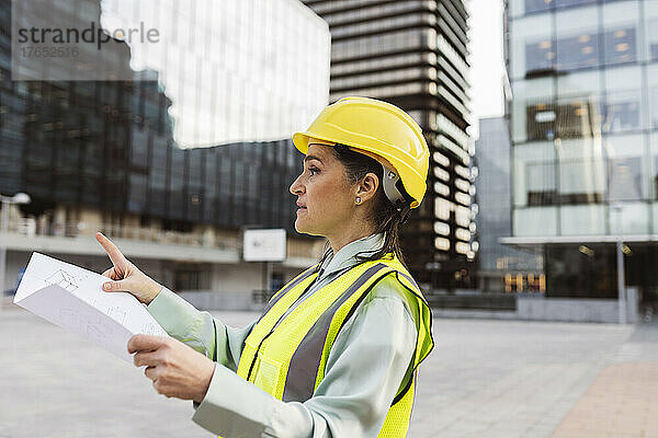 Ingenieur trägt Schutzhelm und hält einen Bauplan in der Hand  der vor einem modernen Gebäude arbeitet