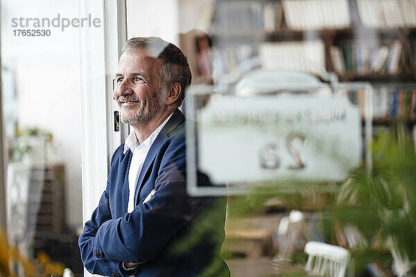 Lächelnder Geschäftsmann mit verschränkten Armen  der durch das Fenster im Café blickt