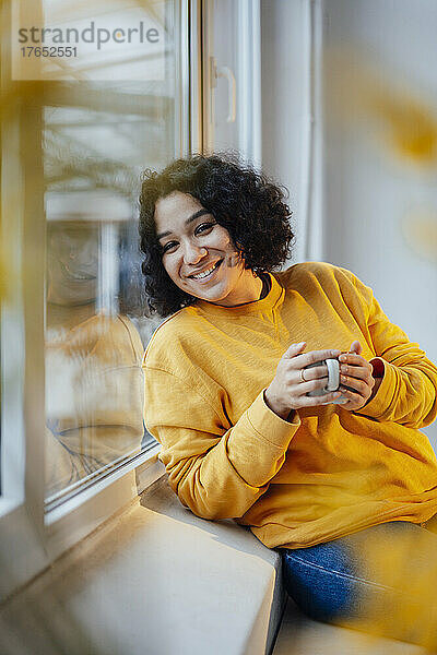 Lächelnde Frau hält eine Kaffeetasse in der Hand und lehnt zu Hause am Fenster