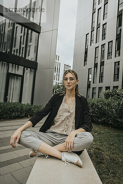 Junge Frau sitzt mit gekreuzten Beinen auf einem Sims vor einem modernen Gebäude