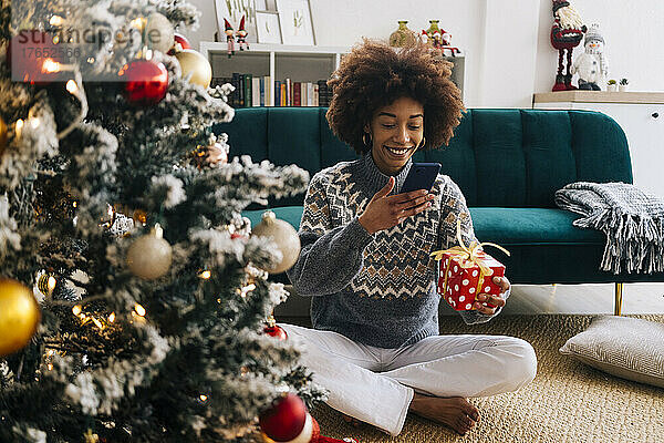 Lächelnde junge Frau fotografiert zu Hause Weihnachtsgeschenk per Handy