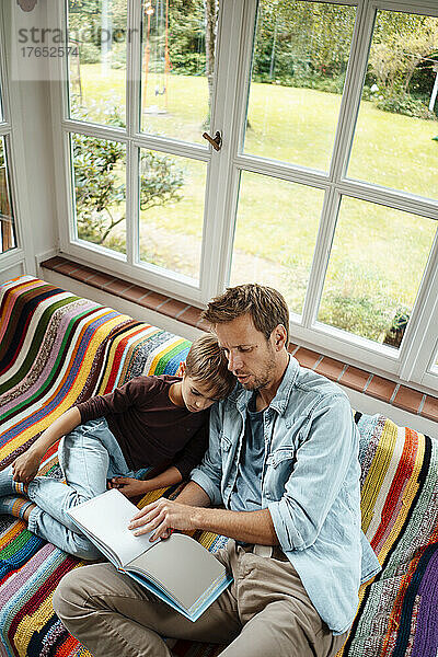 Mann bringt seinem Sohn das Lesen eines Buches bei  der zu Hause auf dem Sofa sitzt