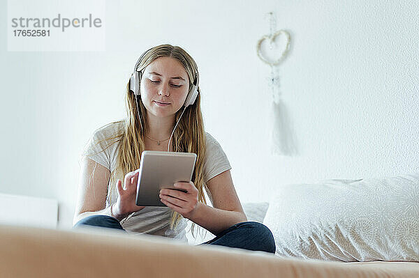 Junge Frau hört Musik über Kopfhörer mit Tablet-PC und sitzt zu Hause im Bett