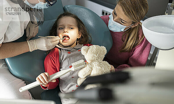 Mutter mit Schutzmaske schaut Tochter an  die vom Zahnarzt in der Klinik untersucht wird