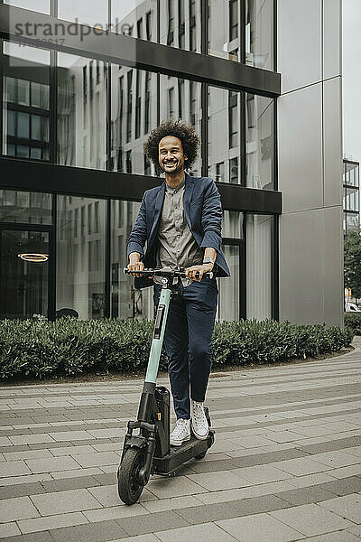 Glücklicher junger Mann fährt mit Elektroroller auf Fußweg vor modernem Gebäude