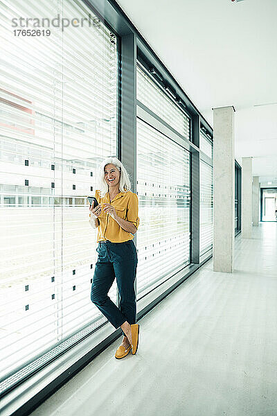 Fröhliche Geschäftsfrau mit Smartphone lehnt an Glaswand im Büro