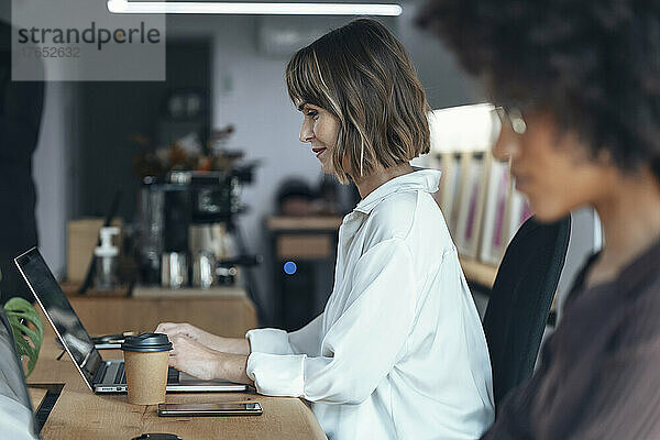 Lächelnde Geschäftsfrau benutzt Laptop und sitzt neben einem Kollegen im Büro