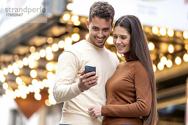 Lächelnder Mann zeigt Freundin Handy vor beleuchteter Decke
