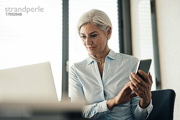 Geschäftsfrau hält Smartphone und schaut im Büro auf Laptop