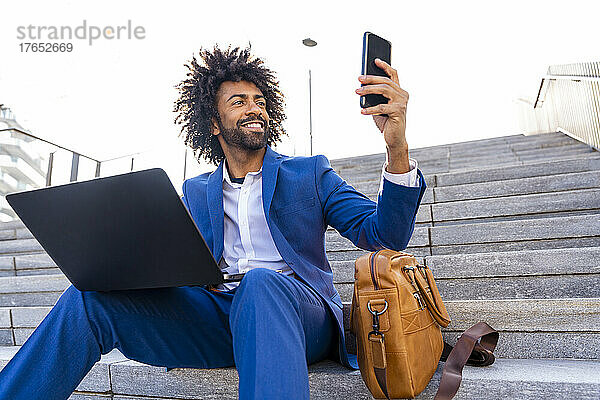 Glücklicher Geschäftsmann mit Laptop  der auf einer Treppe ein Selfie mit dem Mobiltelefon macht