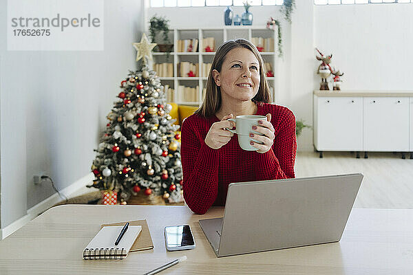 Lächelnde Frau mit Kaffeetasse sitzt am Tisch im Wohnzimmer