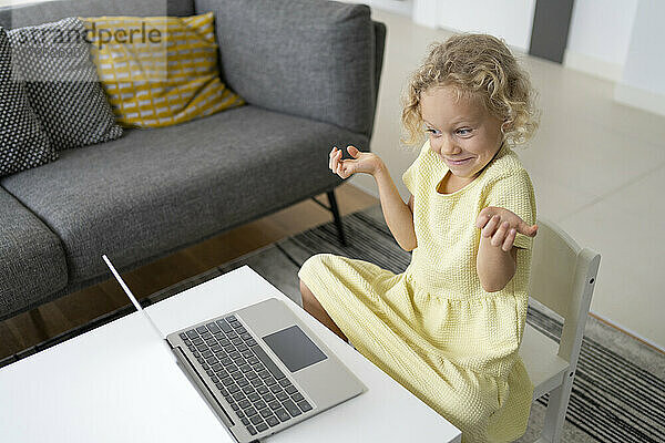 Blond girl gesturing sitting with laptop at table in living room