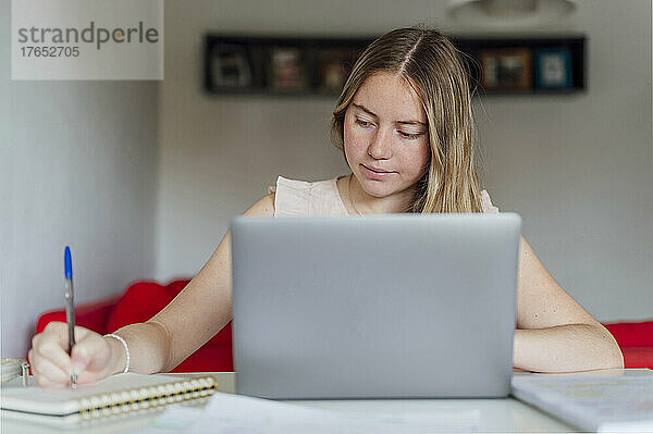 Lächelnde junge Frau schreibt Notizen und sitzt zu Hause am Laptop