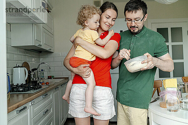 Mann hält Schüssel und steht neben Frau  die ihre Tochter in der Küche trägt