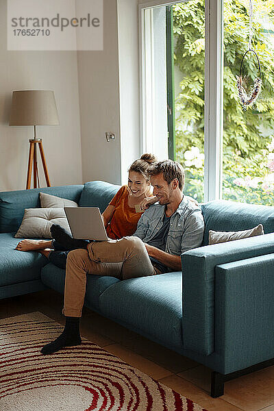 Glückliches Paar teilt sich einen Laptop und sitzt zu Hause auf dem Sofa im Wohnzimmer