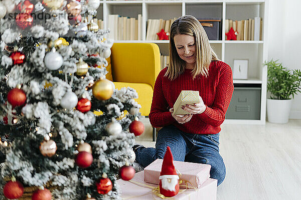 Lächelnde Frau mit Geschenken sitzt zu Hause am Weihnachtsbaum