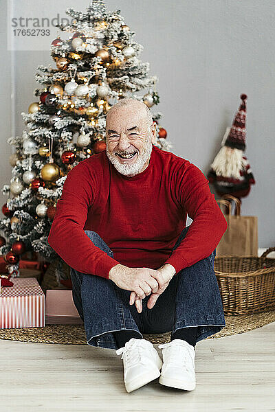 Glücklicher älterer Mann  der zu Hause vor dem Weihnachtsbaum sitzt