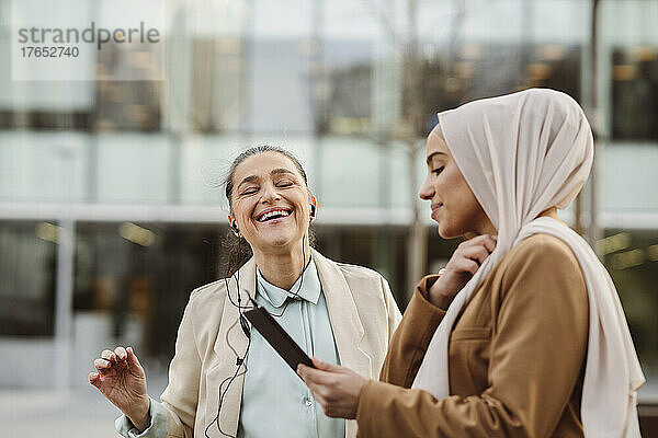 Lächelnde junge Geschäftsfrau mit Tablet-PC steht neben einem glücklichen Kollegen  der In-Ear-Kopfhörer trägt