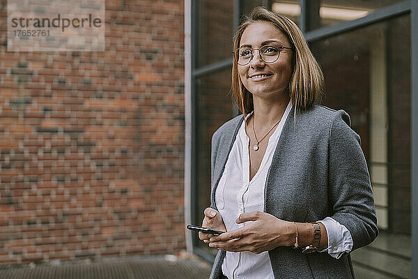 Lächelnde Frau mit Mobiltelefon steht vor dem Gebäude