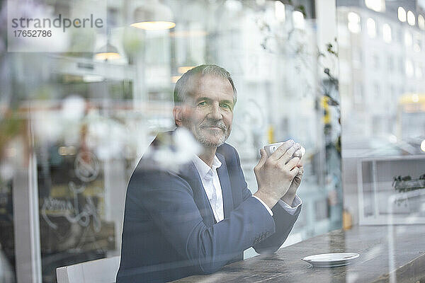 Älterer Geschäftsmann mit Kaffeetasse sitzt am Tisch am Glasfenster