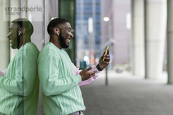 Happy man with wireless in-ear headphones having video call on smart phone by glass wall