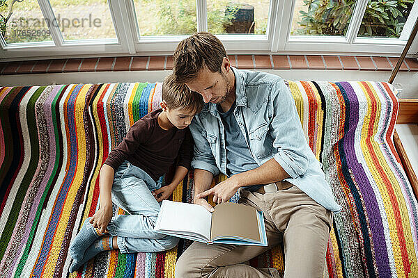 Vater bringt seinem Sohn das Lesen eines Buches auf dem Sofa zu Hause bei