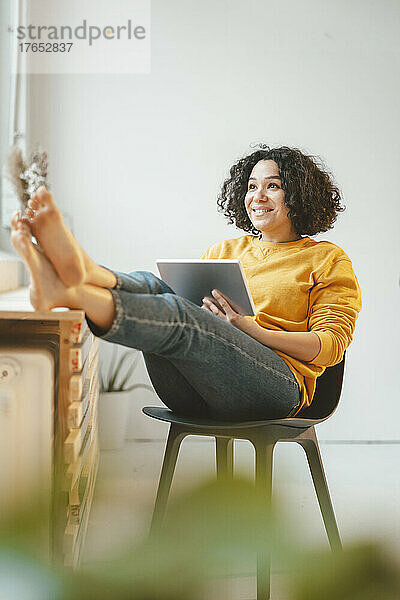 Glückliche schöne Frau  die zu Hause mit Tablet-PC auf einem Stuhl sitzt
