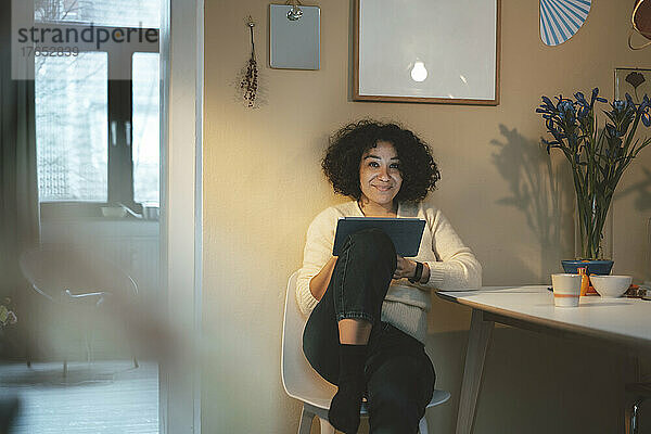 Lächelnde Frau mit Tablet-PC sitzt zu Hause auf einem Stuhl