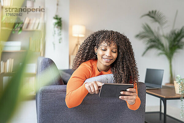 Lächelnde Frau surft mit ihrem Mobiltelefon auf dem Sofa im Wohnzimmer im Internet