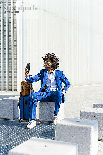 Lächelnder Geschäftsmann mit Gepäck macht ein Selfie per Smartphone und sitzt auf einem Betonblock