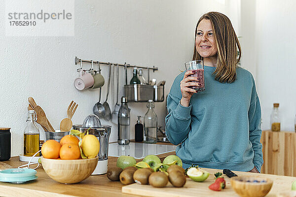 Lächelnde blonde Frau mit einem Glas Smoothie steht am Tisch in der Küche