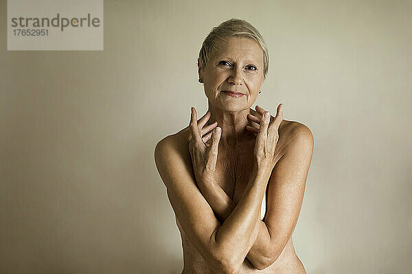 Smiling topless senior woman covering breasts against white background