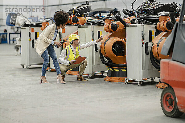 Ingenieur zeigt auf Roboterarm und diskutiert mit Geschäftsfrau  die in der Fabrik einen Tablet-PC hält