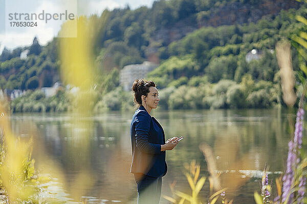 Lächelnde Geschäftsfrau mit Smartphone am Flussufer