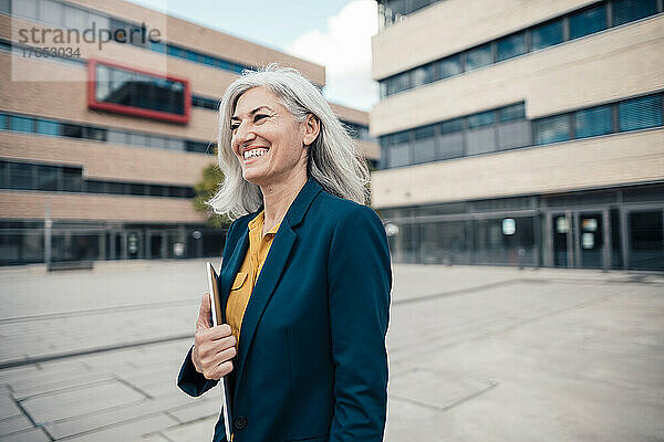 Glückliche Geschäftsfrau mit grauen Haaren hält Laptop im Büropark