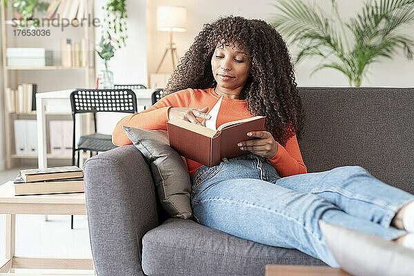 Junge Frau mit lockigem Haar liest ein Buch und sitzt zu Hause auf dem Sofa