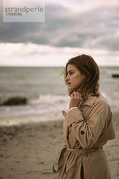 Nachdenkliche junge Frau im Mantel steht bei Sonnenuntergang am Strand