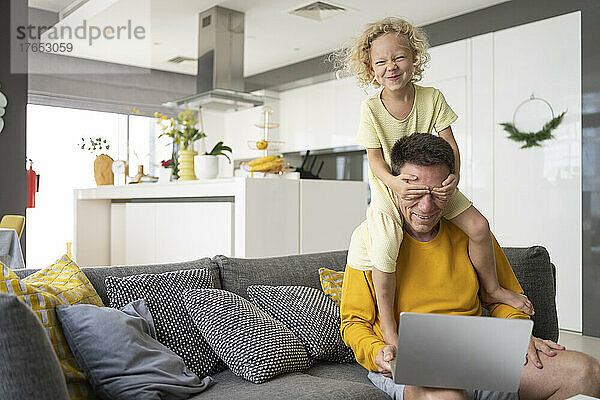 Die schelmische Tochter verdeckt die Augen des Vaters  der zu Hause mit Laptop auf dem Sofa sitzt