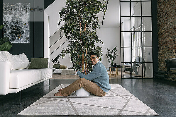 Lächelnder junger Mann mit geschlossenen Augen umarmt Zimmerpflanze  die zu Hause auf dem Teppich sitzt