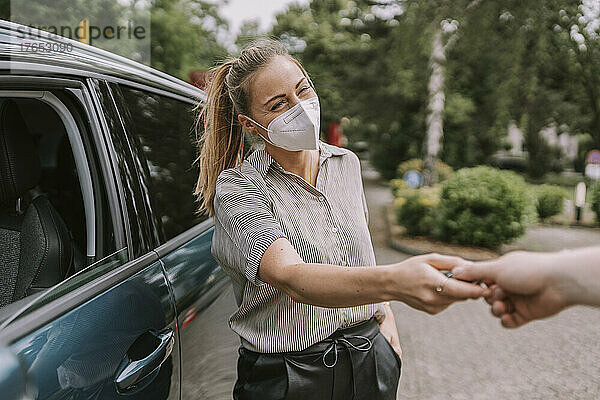 Junge Frau mit schützender Gesichtsmaske gibt einem Freund auf dem Parkplatz den Autoschlüssel